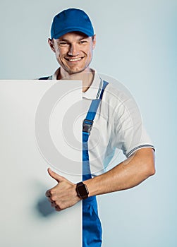 Mockup of smiling young worker man wearing blue uniform. Holding white empty banner in his hand and showing thumb up. Movement