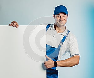 Mockup of smiling young worker man wearing blue uniform. Holding white empty banner in his hand and showing thumb up. Movement