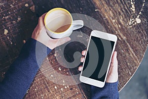 Mockup top view image of woman`s hands holding white mobile phone with blank black screen and a coffee cup on wooden table