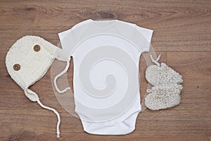 Mockup of white baby bodysuit on wood background with knitted booties and hat. Blank baby clothes template mock up