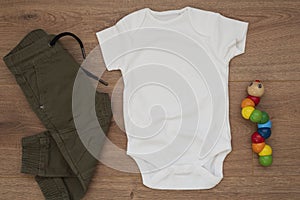 Mockup of white baby bodysuit on wood background with pants and colorful toy. Blank baby clothes template mock up, flat lay styled