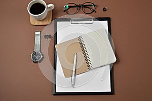 Mockup workspace with blank clip board, office supplies, Clock, pen, coffee cup, notepad and eyeglasses on brown background.
