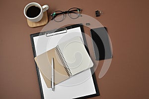 Mockup workspace with blank clip board, office supplies, pen, notepad, smartphone, coffee cup and spectacles on brown background.