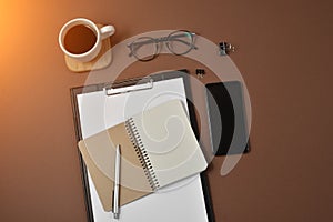 Mockup workspace with blank clip board, office supplies, pen, notepad ,smartphone, coffee cup and spectacles on brown background.