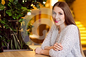 Model with blonde long hair and bright pink lipstick in white sweater sitting on a chair in a cafe with beautiful stylish interior