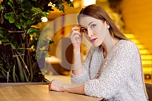 Model with blonde long hair and bright pink lipstick in white sweater sitting on a chair in a cafe with beautiful stylish interior