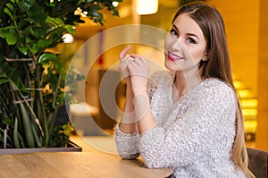 Model with blonde long hair and bright pink lipstick in white sweater sitting on a chair in a cafe with beautiful stylish interior