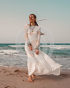 Model in boho style in a white long dress and silver jewelry on the beach. Her hair is braided, and there are many