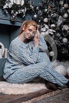 Model of a cute young woman in a stylish knitted blue suit posing on the floor near a beautiful Christmas tree with vintage toys.