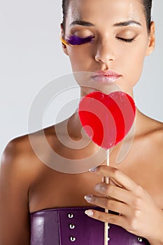 Model with fake bird feather eyelashes holds a heart-shaped lollipop eyes closed