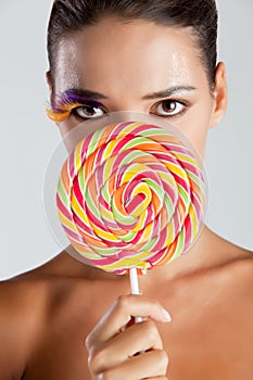 Model with fake bird feather eyelashes holds a rainbow lollipop near her face