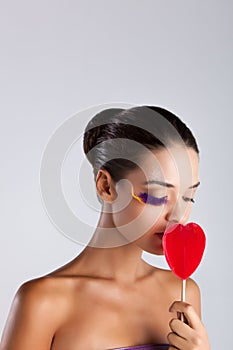 Model with fake feather eyelashes holds a red heart-shaped lollipop near her face