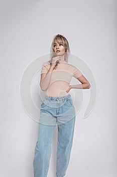 Model of a fashionable young blonde woman in stylish blue jeans in a trendy shirt with a hairstyle stands near a white wall in the