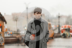 Model of a fashionable young man in an elegant youth checkered coat with a trendy hairstyle stands in the city square with a hot