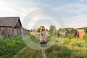 A model girl in a retro dress is holding in her hands a vintage suitcase on an abandoned country road overgrown with grass