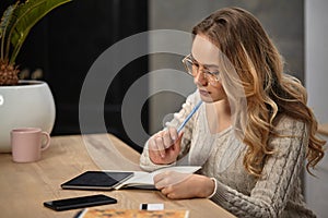 Model in glasses, beige sweater. Sitting at table with cup and palm on it. Writes something in notebook. Tablet