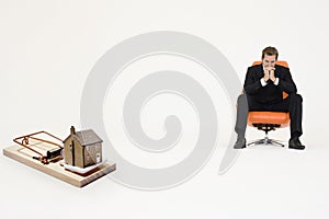 Model home on mouse trap with worried businessman sitting on chair representing increasing real estate rates