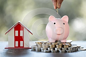 Model of house with female hand putting coin in piggy bank on blurred background. Money saving or investing for home loan or real