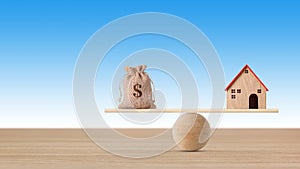 Model house on wooden seesaw balancing with money bag on blue background. Property investment and home mortgage financial real