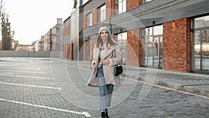 Model of a joyful young woman in fashionable youth outerwear in a knitted stylish hat with a leather handbag travels near a