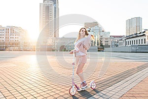 Model in pink dress and pink kick scooter pose against the background of the city landscape in the sunset and smiles