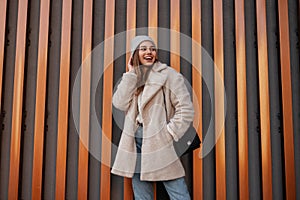 Model of a positive young woman with a beautiful smile in fashionable outerwear in a knitted stylish hat with a leather handbag