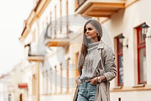 Model of a pretty elegant woman in a chic beige trench coat with brown long hair in a stylish knitted sweater in a jeans walks