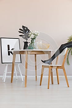 Model`s leg on chair next to wooden table with leaf in dining room interior with poster. Real photo