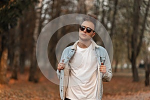 Model trendy American young man in fashionable sunglasses in a stylish denim jacket with a hairstyle in the park. Handsome guy