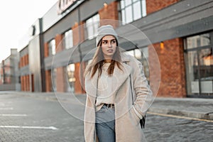 Model of a trendy young woman in fashionable youth outerwear in a knitted stylish hat with a leather handbag posing near a vintage