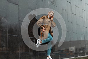 Model of a trendy young woman in a stylish trench coat in blue vintage jeans in fashionable sneakers posing near a black wall in