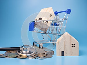 A model wooden house on a shopping cart With a pile of coins and stethoscope On a blue background. Mortgage concept. Money and