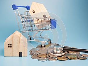 A model wooden house on a shopping cart With a pile of coins and stethoscope On a blue background. Mortgage concept. Money and