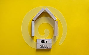 Model of a wooden house. Words `buy, sell` on wooden blocks. Copy space. Business concept. Beautiful yelllow background