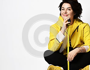 Model in yellow t-shirt, sneakers and jacket, black pants. Showing tape-measure, posing sitting on floor, isolated on white