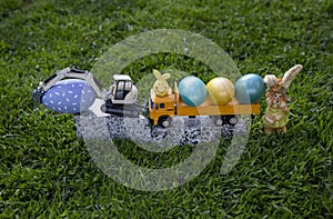 models of a toy excavator and dump truck stand on granite stone in grass along with bright colorful Easter eggs