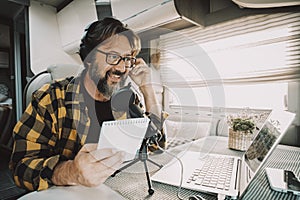 Modern adult man wokring with laptop and microphone inside a camper van in digital nomad job lifestyle. Technology and travel.