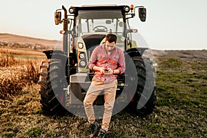 Modern agriculture with technology and machinery concept. Portrait of smiling farmer using smartphone and tractor at harvesting