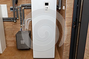 A modern air heat pump installed in the home\'s boiler room, visible plastic pipes and valves.