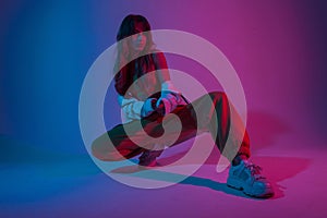 Modern attractive young woman dancer in fashionable youth clothes in a white sneakers sits in a room with bright blue light.