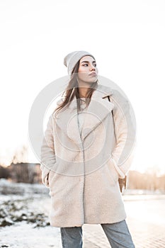 Modern attractive young woman in fashionable winter warm outwear posing in the city on the background of a bright sunset.