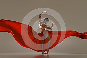 Modern ballet. Handsome young male ballet dancer performing with red silk fabric against grey studio background