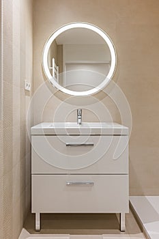 Modern bathroom with beige tiles, furniture and round large mirror with lighting