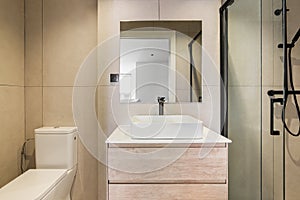 Modern bathroom with beige tiles. Square mirror, toilet, white sink and shower zone with glass partition.