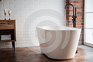 Modern bathroom with freestanding white acrylic bathtub and black loft mixer. Walls made of bricks, parquet is wooden on floor,