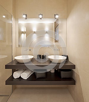 Modern bathroom front view with large light marble slabs. There are two sinks with a large mirror illuminated by spotlights
