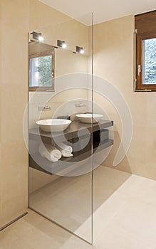 Modern bathroom front view with large light marble slabs. There are two sinks with a large mirror illuminated by spotlights