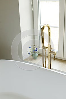 Modern bathroom with stylish golden faucet, white bathtub and travertine tiles. Stylish bathroom details with summer flowers