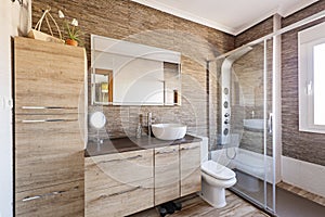 Modern bathroom with wooden furniture with large drawers, walls with similar stone tiles and a shower cabin with jets and music