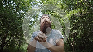 A modern bearded man in a cap trains in the forest and kneads joints